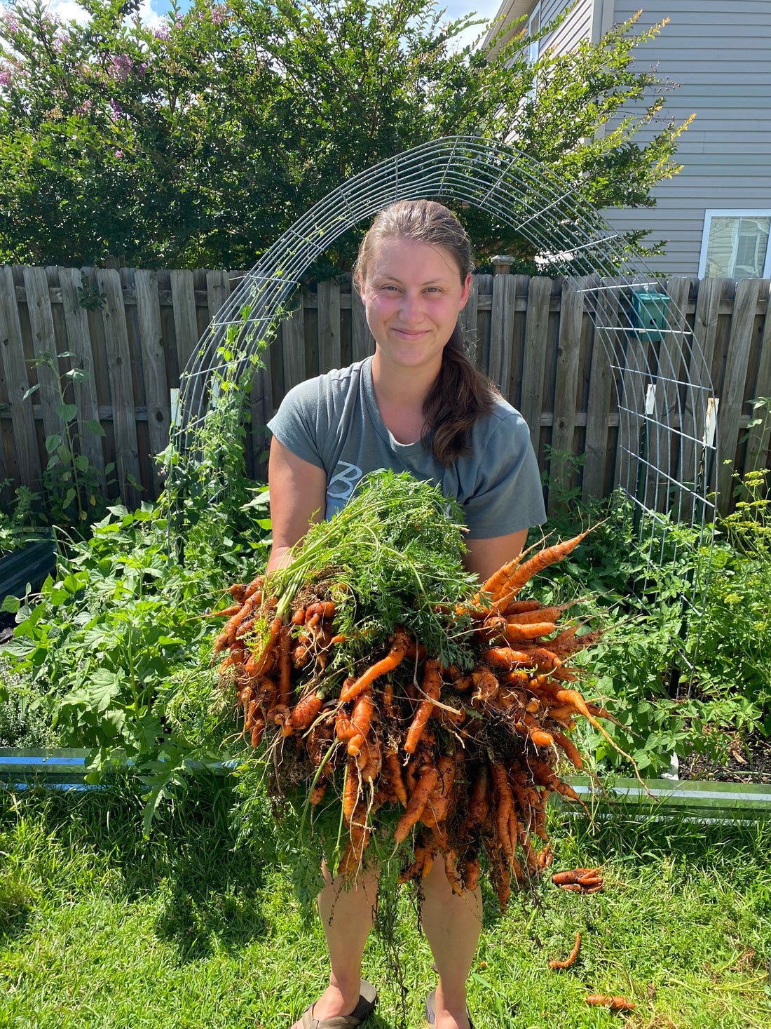 Raven, owner of Growth and Gardening, standing in her garden holding two large hand fulls of carrots she grew